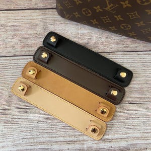 May include: Four rectangular leather handle covers arranged diagonally, featuring gold-tone metal snap buttons at each end. The covers are in shades of black, dark brown, tan, and light beige, positioned below a brown monogrammed handbag.