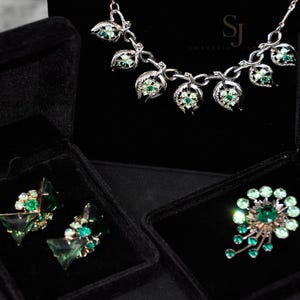 Pode incluir: Um conjunto de joias prateadas com um colar, brincos e um broche, todos adornados com pedras preciosas verdes. O colar e o broche têm um design floral, enquanto os brincos têm uma forma geométrica. As joias são exibidas em caixas de veludo preto.