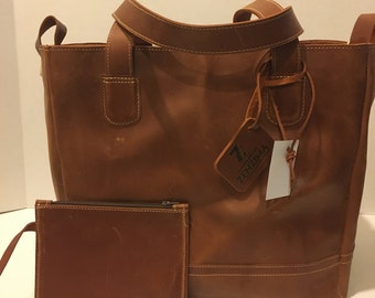 May include: A brown leather tote bag with a smaller matching leather pouch. The tote bag has a top handle and a detachable shoulder strap. The bag has a small leather tag with the text "ZenZima" on it.