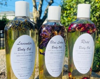Op de afbeelding: Drie glazen flessen met lichaams olie met etiketten die "Lavender Body Oil", "Blue Lotus Body Oil" en "Rose Body Oil" zeggen. Elke fles bevat gedroogde bloemen en is geëtiketteerd als 100% puur en natuurlijk.
