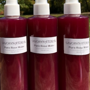 May include: Three glass bottles with white spray tops filled with a deep red liquid. Each bottle has a white label with the text "SAVORYNATURAL Pure Rose Water Find yourself @SavoryNatural.com Here for you!"