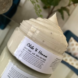 May include: A clear plastic jar filled with a thick, off-white hair mask cream that has a swirled, peaked top. The white label features black text reading Chebe & Henna HAIR MASK with IRISH SEA MOSS, GROWTH SUPPORT & MOISTURE RETENTION, and directions.