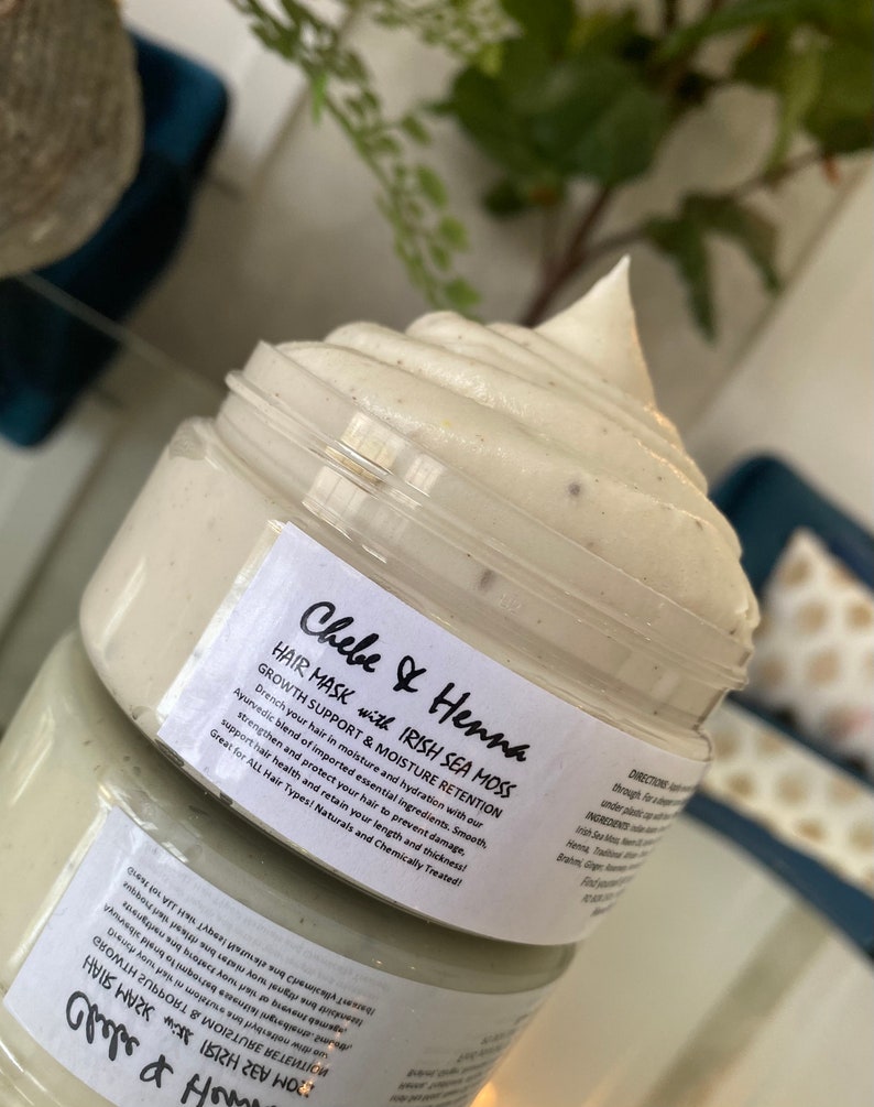 May include: A clear plastic jar filled with a thick, off-white hair mask cream that has a swirled, peaked top. The white label features black text reading Chebe & Henna HAIR MASK with IRISH SEA MOSS, GROWTH SUPPORT & MOISTURE RETENTION, and directions.