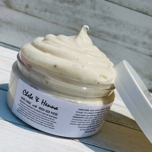 May include: A round plastic jar filled with a thick, off-white hair mask featuring small dark particles. The white label reads Chebe & Henna Hair Mask with Irish Sea Moss, Growth Support & Moisture Retention, along with directions and ingredients.