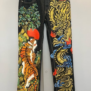 Op de afbeelding: Een donkerblauwe spijkerbroek met een handgeschilderd ontwerp. Het linkerbeen heeft een tijger met oranje en zwarte strepen, omringd door groen gebladerte en een rode zon. Het rechterbeen heeft een gouden draak met blauwe en rode accenten, omringd door wolken.