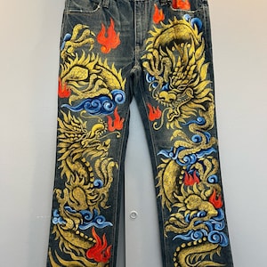 Puede incluir: Un par de pantalones vaqueros azules con un diseño pintado a mano de un dragón dorado con llamas rojas y nubes azules. Los vaqueros son de estilo bootcut.