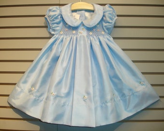 Peut inclure: Une robe de bébé bleu clair avec un col blanc et des fleurs brodées. La robe a un corsage smocké et une jupe ample.