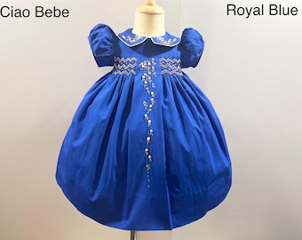 May include: A royal blue smocked dress with a white collar and embroidered flowers. The dress has puffed sleeves and a full skirt. The text "Ciao Bebe" and "Royal Blue" are visible in the image.