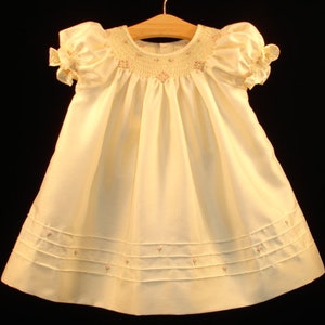 May include: A white satin baby dress with a smocked bodice and embroidered floral details. The dress has puffed sleeves with lace trim and a gathered skirt.