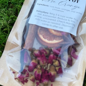 May include: A clear plastic bag containing a simmer pot blend. The label reads "MANESS FAMILY FARM SIMMER POT Rose Orange." Ingredients include rose buds, orange slices, and cinnamon sticks. The bag is filled with dried rose buds and other botanicals.