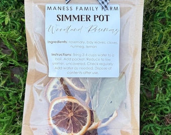 May include: A clear plastic bag containing a simmer pot kit, with the text "MANESS FAMILY FARM SIMMER POT Woodland Rosemary." The bag includes dried lemon slices, bay leaves, and spices. A black and white checkered bow adorns the top of the bag.