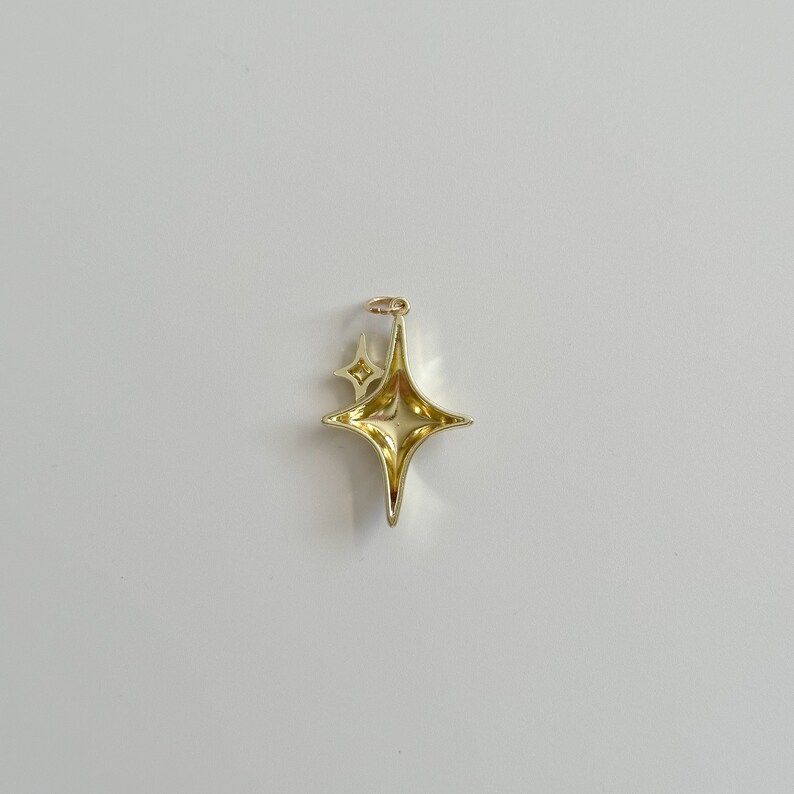 Peut inclure: Pendentif dor&eacute; en forme d'&eacute;toile &agrave; quatre branches avec une petite &eacute;toile &agrave; c&ocirc;t&eacute;. Le pendentif est dot&eacute; d'une boucle en haut pour une cha&icirc;ne. L'&eacute;toile a une surface brillante et r&eacute;fl&eacute;chissante.