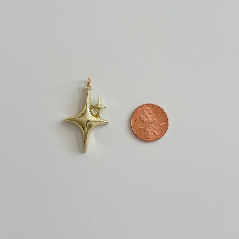 Peut inclure: Un pendentif en forme d'&eacute;toile dor&eacute;e avec une petite &eacute;toile attach&eacute;e. Le pendentif est brillant et r&eacute;fl&eacute;chissant. Une pi&egrave;ce de monnaie est &agrave; c&ocirc;t&eacute; du pendentif pour la comparaison de taille. Le pendentif est probablement pour les bijoux.