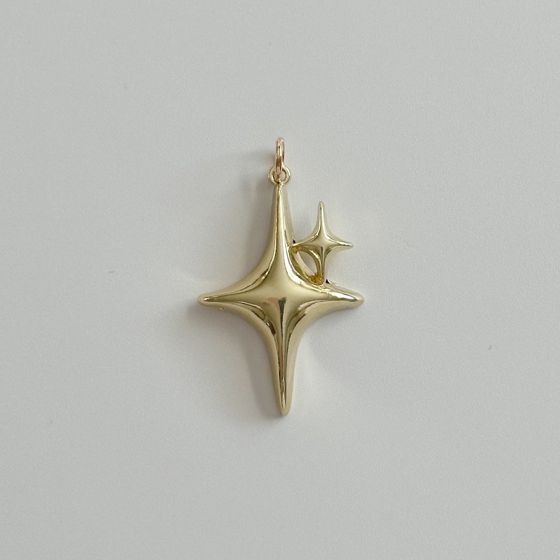 Peut inclure: Un pendentif dor&eacute; en forme d'&eacute;clat d'&eacute;toile. Le pendentif a une forme d'&eacute;toile plus grande avec une forme d'&eacute;toile plus petite attach&eacute;e. Le pendentif a une petite boucle en haut pour une cha&icirc;ne. Le pendentif est en m&eacute;tal brillant et r&eacute;fl&eacute;chissant.