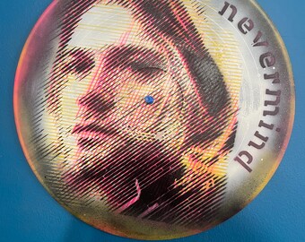 May include: A round, colorful artwork featuring a portrait with pink, yellow, and black tones. The word "nevermind" is printed in a semi-circle around the portrait. A small blue gem is in the center of the artwork.
