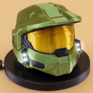 Puede incluir: Un casco verde y amarillo, que recuerda a un personaje de videojuego, se encuentra sobre una base circular negra. El casco tiene una visera y luces a ambos lados. Un cable negro se extiende desde la base.