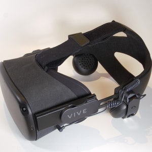 May include: A black VR headset with a black strap and a black headband. The headset has a black logo that says "VIVE" on the side.