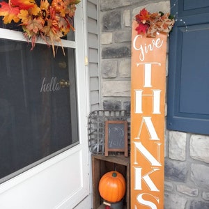 May include: An orange wooden sign with the words "GIVE THANKS" in white lettering stands next to a door. A fall wreath hangs on the door. A pumpkin and chalkboard are displayed in a wooden crate.