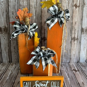 May include: Orange wooden pumpkin decorations with black and white checkered bows, faux leaves, and twine accents. A small rectangular sign with the words "FALL SWEET FALL" is in front of the pumpkins. The pumpkins are various sizes and shapes.