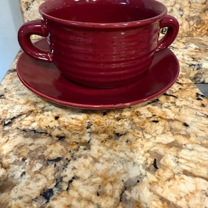 May include: A deep red ceramic soup bowl with two handles, resting on a matching saucer. The bowl and saucer have a textured, slightly rustic appearance. The set is designed for serving soup or other warm dishes.