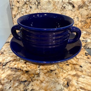 May include: A deep blue ceramic soup bowl with two handles, resting on a matching saucer. The bowl has a ribbed design and a glossy finish. This set is ideal for serving soup or other dishes.