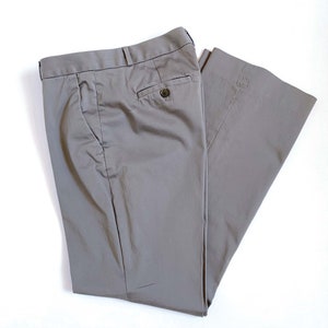 May include: A pair of gray dress pants, folded neatly on a white surface. The pants have a button closure, belt loops, and pockets. The fabric appears to be a smooth, woven material. The pants are a neutral color, suitable for various outfits.