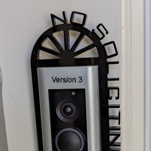 May include: A black and silver Ring doorbell with the text "Version 3" and "ring" on the front. The doorbell is mounted on a white wall with a black decorative mount.