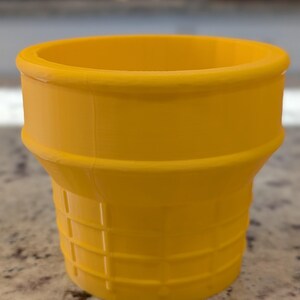 May include: A yellow plastic ice cream cone shaped cup. The cup has a waffle cone texture.
