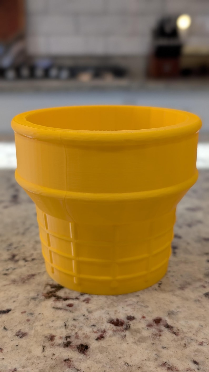May include: A yellow plastic ice cream cone shaped cup. The cup has a waffle cone texture.