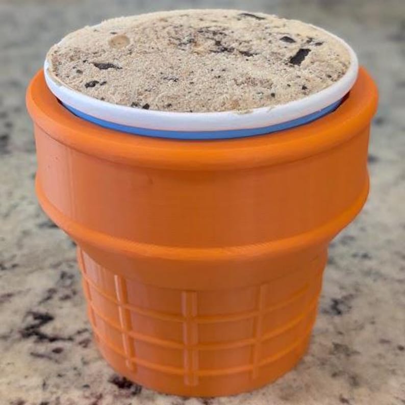 May include: Orange plastic ice cream cone mold with a white and blue lid. The mold is filled with a light brown ice cream with chocolate chips.