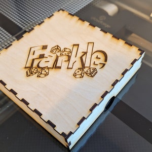 May include: A wooden box with a laser-engraved design of the word "Farkle" and dice. The box is light brown and has a dark brown border.