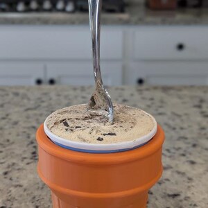 May include: A close-up of a single-serving ice cream cone with a silver spoon sticking out of the top. The ice cream is a light brown color and has chocolate chips in it. The cone is orange and has a waffle texture.