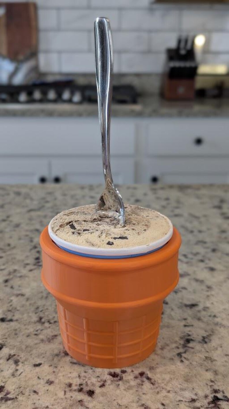 May include: A close-up of a single-serving ice cream cone with a silver spoon sticking out of the top. The ice cream is a light brown color and has chocolate chips in it. The cone is orange and has a waffle texture.