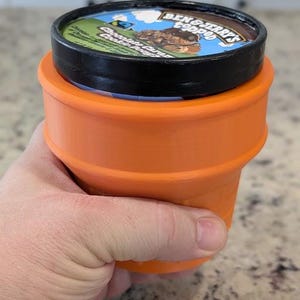 May include: A black and orange ice cream container with a Ben & Jerry's label that reads "Chocolate Caramel Cookie Dough" and a cartoon image of a bear. The container is sitting in an orange silicone ice cream holder.
