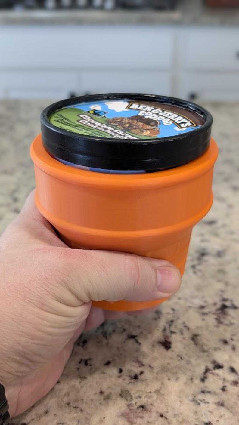 May include: A black and orange ice cream container with a Ben & Jerry's label that reads "Chocolate Caramel Cookie Dough" and a cartoon image of a bear. The container is sitting in an orange silicone ice cream holder.