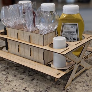 Może przedstawiać: Drewniany kosz w stylu stołu piknikowego mieści przezroczyste plastikowe sztućce, butelki z przyprawami i butelkę musztardy. Kosz jest wykonany z jasnego drewna i ma mały, biały świecznik.