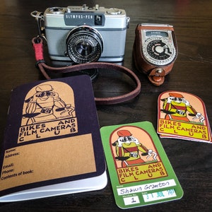 May include: A black and brown membership card for the Bikes and Film Cameras Club. The card has the club's logo and the text "Bikes and Film Cameras Club". The card also has the member's name, membership number, and date. The card is on a wooden table next to a vintage camera and a light meter.