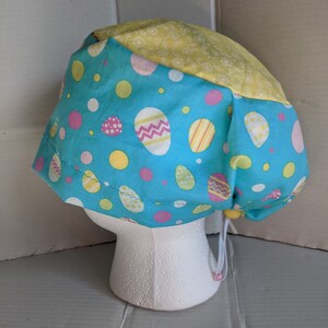 May include: A blue and yellow scrub hat with an Easter egg pattern. The hat has a tie at the back.