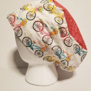 May include: A white scrub hat with a red and white patterned fabric on the side. The hat features a repeating pattern of bicycles with flowers in baskets.