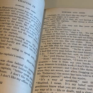 May include: Open book with text on aged paper. The left page displays "CHAPTER IX" and "POWDER AND ARMS." The right page continues the text. The book's pages are yellowed, suggesting age. The text is in black.