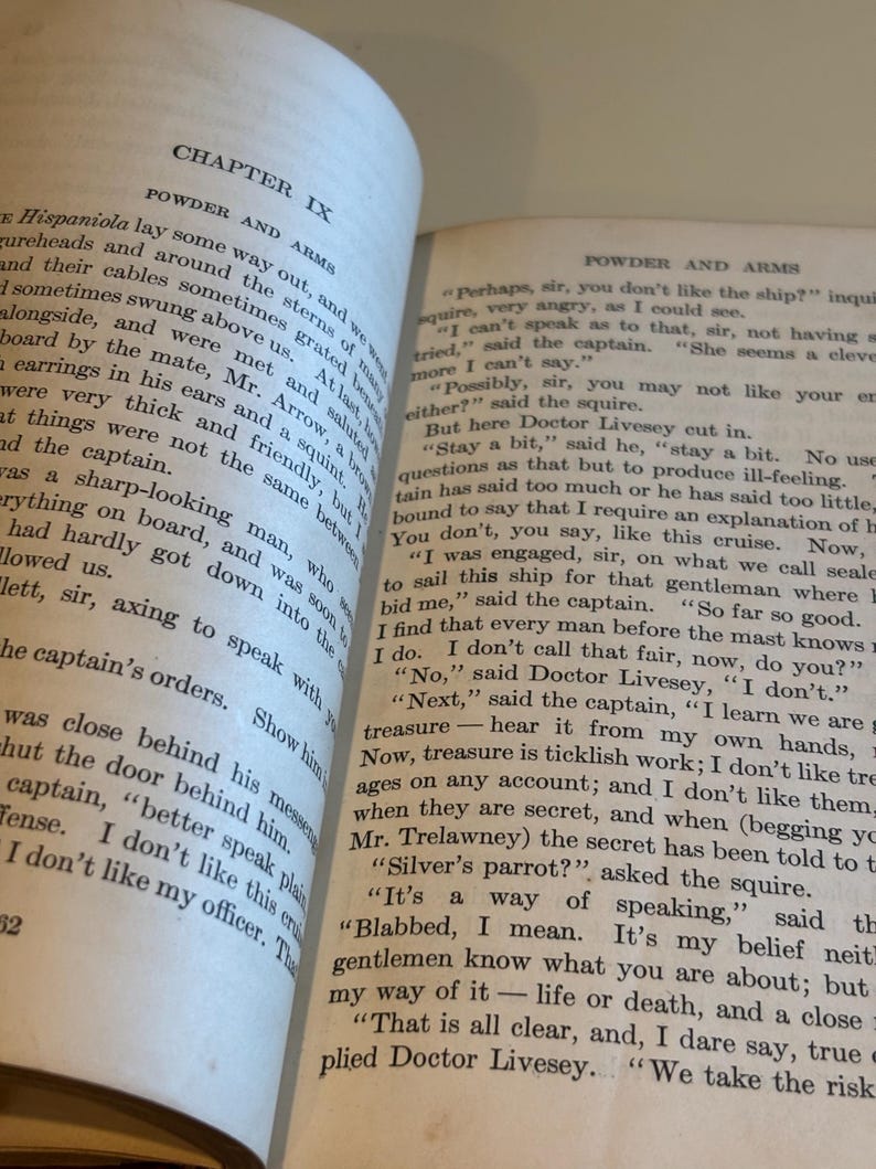 May include: Open book with text on aged paper. The left page displays "CHAPTER IX" and "POWDER AND ARMS." The right page continues the text. The book's pages are yellowed, suggesting age. The text is in black.