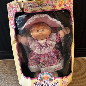 Könnte beinhalten: Eine Cabbage Patch Kids 10th Anniversary Edition Puppe in ihrer Originalverpackung. Die Puppe hat blondes Haar, blaue Augen und trägt ein Blumenkleid mit Spitzenbesatz sowie eine rosa Haube. Der Karton ist mit Blumengrafiken verziert.