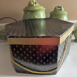May include: Hexagonal black box with gold fleur-de-lis pattern and gold trim. The box has a gold rope handle and is sitting on a white surface. Other green ceramic containers are in the background.