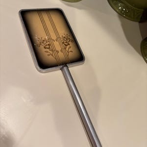 May include: A vintage hand mirror with a silver handle and a rectangular, gold-toned frame. The frame has a floral design with brown lines and a brown background.