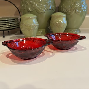 May include: Two ruby red glass bowls with handles. The bowls have a textured, ribbed design and are set on a white surface. Other items in the background include green ceramic jars and a stack of green plates.