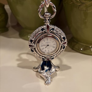 Può includere: Un orologio al quarzo argentato su un supporto decorativo. Il quadrante dell'orologio presenta numeri romani. Il supporto e la cassa presentano dettagli intricati con accenti smaltati blu e rossi. L'orologio è progettato per essere posizionato su una superficie.