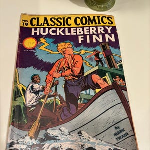 Puede incluir: Portada del número 19 de Classic Comics con Huckleberry Finn de Mark Twain. El arte de la portada representa a Huckleberry Finn en un bote con Jim, remando por un río con rayos en el fondo.