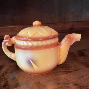 May include: A yellow ceramic teapot with a brown lid and a camel-shaped spout. The teapot has a decorative design of leaves and a rope-like pattern around the middle.