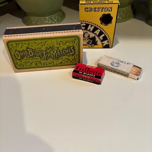 Może przedstawiać: Zbiór vintage pudełek zawierających zapałki, ostrza do golenia i kredę. Pudełko zapałek jest zielone i złote z tekstem "Ohio Blue Tip Matches" i "Strike Anywhere". Pudełko z ostrzami do golenia jest czerwone i czarne z tekstem "Thin Gillette 10 Blades". Pudełko z kredą jest żółte z tekstem "Creston Chalk" i czarnym kółkiem.