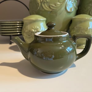 Może przedstawiać: Mały, oliwkowozielony ceramiczny imbryk z pokrywką i uchwytem. Czajniczek ma błyszczące wykończenie i białą obwódkę. Za imbrykiem znajduje się kilka zielonych elementów ceramicznych, w tym stos talerzy.